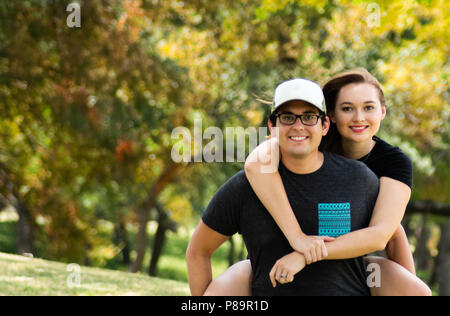 Giovane uomo che porta attraente giovane donna piggyback con colorate Foglie di autunno a distanza Foto Stock