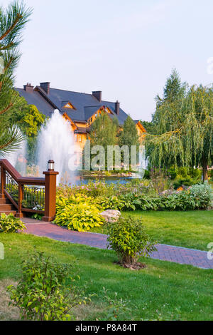 Fontane in lago di fronte un grande cottage di legno circondato da un parco verde, prato e alberi con una luce blu del cielo in background. residence Foto Stock