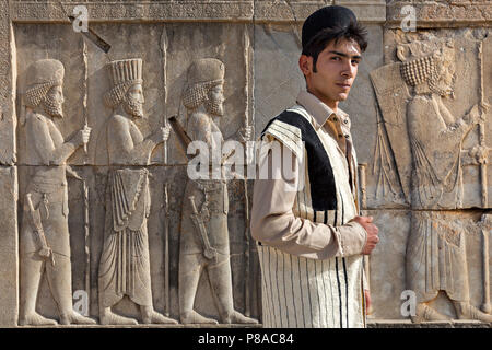 Giovane bakhtiari etnico nei suoi abiti tradizionali con rilievi persiani dietro di lui, a Persepolis, Iran. Foto Stock