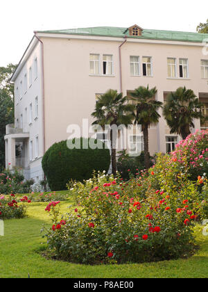L'angolo dell'edificio bianco con un tetto verde e vicino a crescere alberi di palma. Circondato da un letto di fiori con rose rosse . Per il vostro design Foto Stock