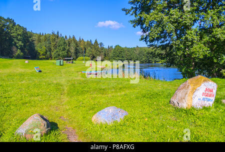 Una soleggiata radura sulla riva di un lago tranquillo o stagno con un piccolo molo per la pesca e un gazebo circondato da alberi di alto fusto. luogo di riposo, pesca e pic Foto Stock