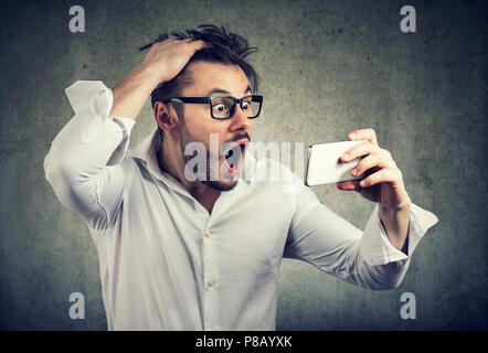 Crazy giovane uomo avente perdita di capelli e guardare lo smartphone nel grande espressione di stupore urlando su sfondo grigio Foto Stock