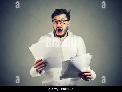 Giovane ragazzo scioccato in bicchieri guardando papers documenti bancari con la lista delle fatture e dei canoni e la sensazione di essere sottolineato su sfondo grigio Foto Stock