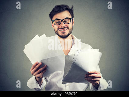 Felice giovane uomo in bicchieri guardando attraverso i documenti cartacei con buoni risultati e sorridente su sfondo grigio Foto Stock