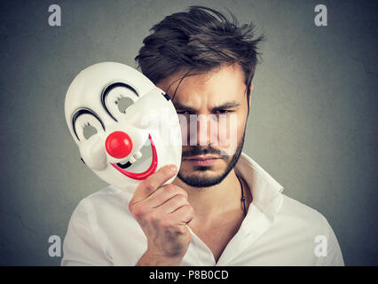 Giovane uomo barbuto holding maschera felice e la sensazione di essere triste e deprimente mentre guardando la fotocamera su sfondo grigio Foto Stock