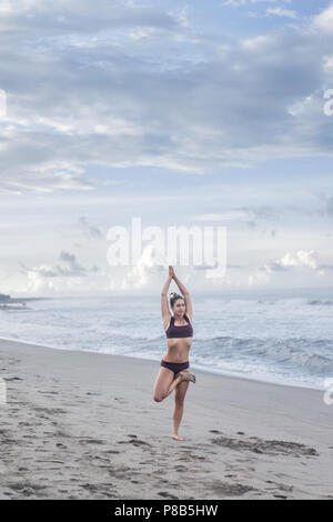 Attraente giovane donna a praticare yoga in posa ad albero (Vrksasana) sulla riva del mare Foto Stock