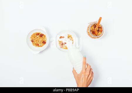 Donna facendo sano muesli dado prima colazione per due, vista dall'alto. La femmina lato versare il latte in un recipiente riempito con fatti in casa oat muesli, laici piatta su bianco b Foto Stock