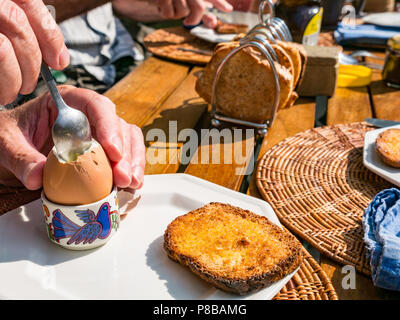 Uomo che mangia uova sode in una tazza di uova con toast sul tavolo esterno in legno da giardino con toast rack al sole a colazione Foto Stock
