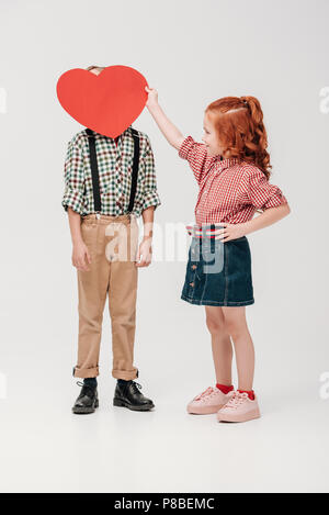 Little Girl holding cuore rosso simbolo vicino alla faccia del ragazzino isolato su grigio Foto Stock