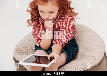 Focalizzato redhead piccolo bambino seduto e con tavoletta digitale isolato su grigio Foto Stock