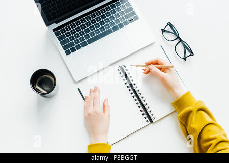 Vista dall'alto di mani femminili matita di contenimento a tavola con laptop, occhiali e la tazza di caffè Foto Stock