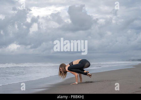 Montare la giovane donna a praticare yoga in Crow pongono (Bakasana) sulla riva del mare Foto Stock