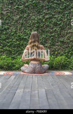 Vista posteriore della donna a praticare yoga in lotus pongono rendendo namaste gesto dietro alla schiena di fronte a parete ricoperta con foglie di colore verde Foto Stock