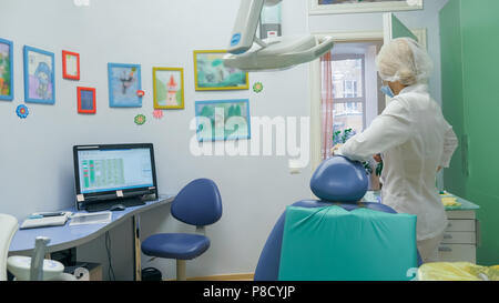 Bambino con una madre a un dentista di ricezione. La ragazza si trova in cattedra, dietro la madre. Il dottore lavora con un assistente. Procedura per la perforazione di un dente. Impostazione di una sigillatura temporanea. Foto Stock