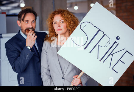 Imprenditrice azienda grande poster con la parola sciopero. Dai capelli rossi che mostra fwmale infelice emozioni, uomo felice in piedi vicino a lei di mangiare la torta. Tonica conc Foto Stock