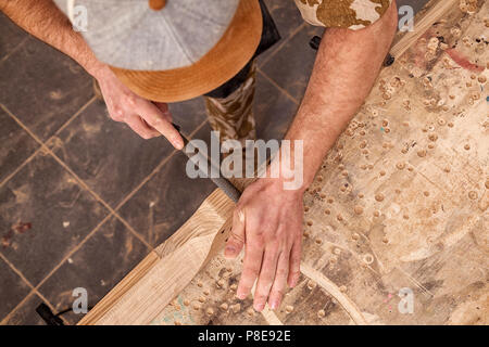 Chiusura del giovane uomo builder indossa in un plaid shirt il trattamento di un prodotto di legno con uno scalpello in officina, close-up Foto Stock