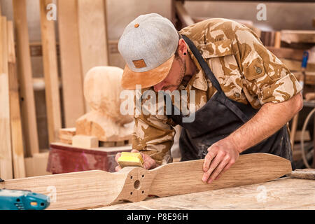 Un uomo di lavoro in un cappuccio e camicia lucida il blocco di legno con carta vetrata prima della verniciatura in officina, in background, strumenti e mach di perforazione Foto Stock