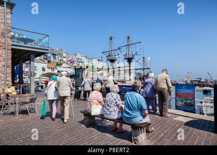 23 Maggio 2018: Brixham, Devon, Regno Unito - i cittadini anziani rilassante sul lungomare a Brixham Harbour, con la replica di Golden Hind nave a vela. Foto Stock