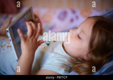 Poco Caucasian due-anno-vecchia ragazza distesa su un plaid cuscino e guardando cartoni animati su un computer tablet. Foto Stock