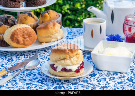Il tè del pomeriggio con torte e Inglese tradizionale con scones con confettura di fragole e clotted cream impostato su un tavolo in giardino. Sala da pranzo all'aperto. Foto Stock