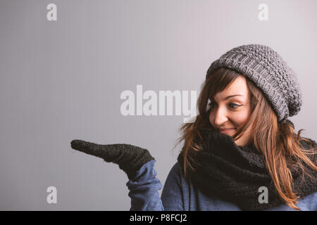 Sorridente giovane donna in inverno la moda tenendo fuori il suo vuoto palm guanti con copia spazio per la collocazione di prodotti su un grigio di sfondo per studio Foto Stock