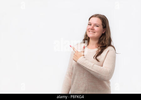 Felice caucasian giovane donna in maglione sorridente ampio alla fotocamera, puntando le dita lontano, mostrando qualcosa di interessante ed eccitante sulla parete di studio. Co Foto Stock