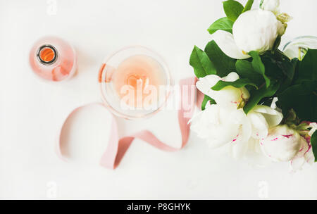 Flat-lay di vino rosato in vetro e bottiglia, rosa nastro decorativo, peonia Fiori su fondo bianco, vista dall'alto, composizione orizzontale. Estate celeb Foto Stock