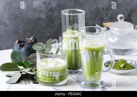 Tè verde Matcha iced latte macchiato o un cocktail in tre diversi tipi di bicchieri con cubetti di ghiaccio, matcha polvere e il contenitore del latte su bianco tavola di marmo, decorata da g Foto Stock