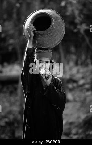 Una ragazza del Kashmir mangia un Apple mentre si trasporta un grande boccale acqua sulla sua testa - KASHMIR INDIA Foto Stock