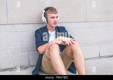 Annoiato teen seduto a terra contro una parete mentre si ascolta la musica attraverso le cuffie. Foto Stock