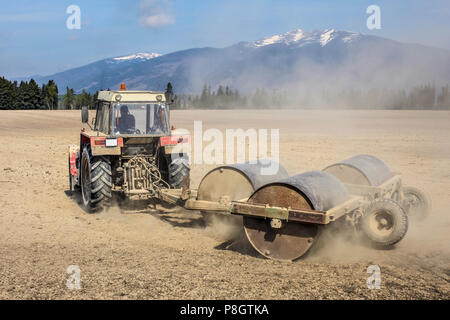 Il trattore il traino di pesanti rulli metallici a secco su un campo con le montagne sullo sfondo. Molla la preparazione di massa. Foto Stock