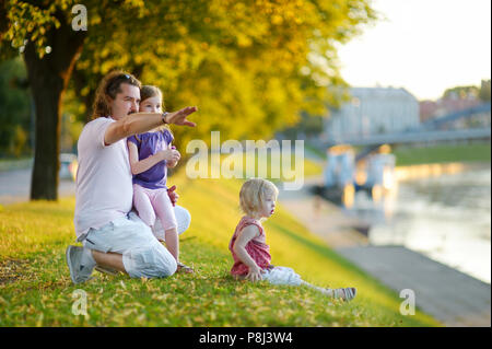 Giovane padre e le sue due piccole figlie seduta da un fiume e guardare le canoe passando da Foto Stock