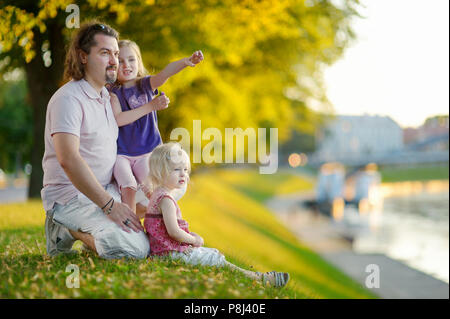 Giovane padre e le sue due piccole figlie seduta da un fiume e guardare le canoe passando da Foto Stock