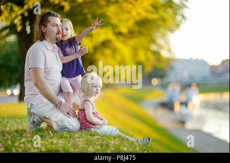 Giovane padre e le sue due piccole figlie seduta da un fiume e guardare le canoe passando da Foto Stock