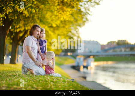 Giovane padre e le sue due piccole figlie seduta da un fiume e guardare le canoe passando da Foto Stock