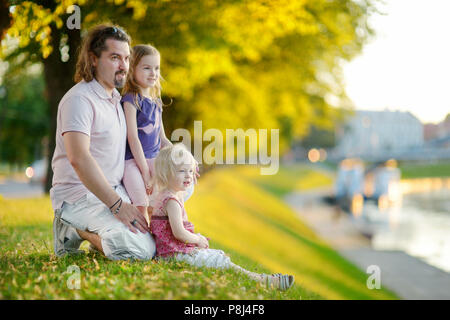 Giovane padre e le sue due piccole figlie seduta da un fiume e guardare le canoe passando da Foto Stock