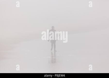 Trattativa, NEW JERSEY - Aprile 28, 2018: un surfista passeggiate lungo la spiaggia su una molto nebbiosa mattina di primavera Foto Stock