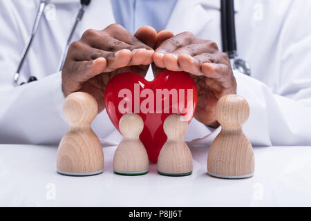 Medico la mano che copre la pedina di legno Famiglia e cuore rosso su bianco scrivania Foto Stock