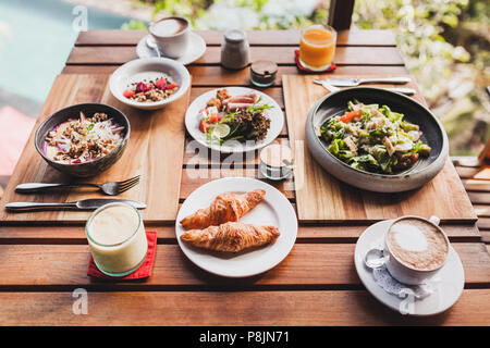 La prima colazione per due in un tavolo di legno. Croissant, insalata fresca, succo naturale, caffè, yogurt con muesli e frutta Foto Stock