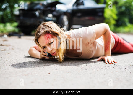 Giovane donna ferita giacente sulla strada dopo un incidente di automobile, facendo una telefonata. Foto Stock