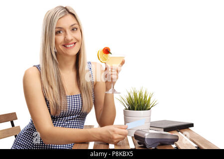 Giovane donna tenendo un cocktail e una carta di credito seduti ad un tavolo isolato su sfondo bianco Foto Stock