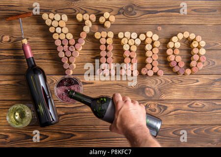 Maschio lato versando il vino in bicchieri contro il vino di scritte fatte di tappi contro lo sfondo di legno Foto Stock