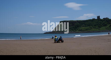 I vacanzieri seduti da, guardando le onde e passando per una racchetta in mare nel North Yorkshire holiday resort di Scarborough,UK sulla baia del nord Foto Stock