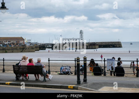 Le famiglie e le persone su un banco e altri sulla spiaggia sabbiosa a Scarborough,UK con il porto e il faro in background Foto Stock