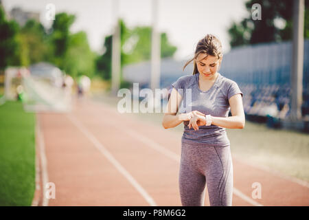 Sport e tecnologia. bellissima giovane donna whiteskinned con coda di cavallo alla esecuzione di stadium di fronte di allenamento utilizza una scattante Smart clock sul suo braccio per Foto Stock