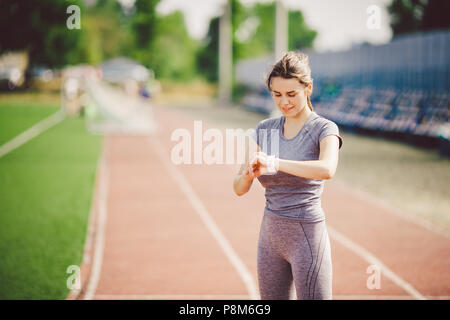 Sport e tecnologia. bellissima giovane donna whiteskinned con coda di cavallo alla esecuzione di stadium di fronte di allenamento utilizza una scattante Smart clock sul suo braccio per Foto Stock