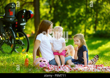 Giovane madre e le sue figlie avente un picnic nel parco sulla bellissima giornata estiva Foto Stock