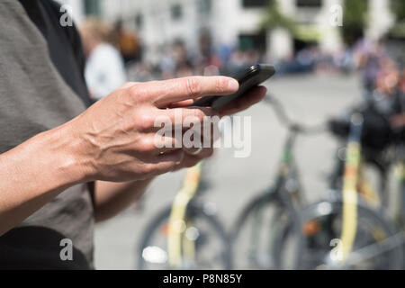 Primo piano di un giovane uomo caucasico utilizzando uno smartphone sulla strada, accanto ad alcune biciclette parcheggiate Foto Stock