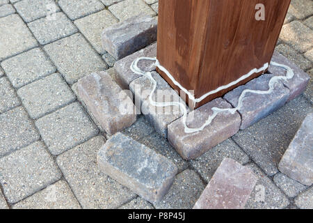 Colla applicata a mattoni grigi attorno ad un pilastro di legno durante l'installazione di un nuovo gazebo sul patio Foto Stock