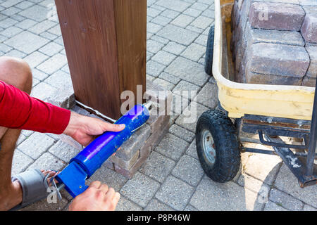 L'uomo l'applicazione di colla da una pistola per colla per muratura come egli stabilisce un pilastro intorno a una nuova gamba di legno su un gazebo sul suo patio Foto Stock